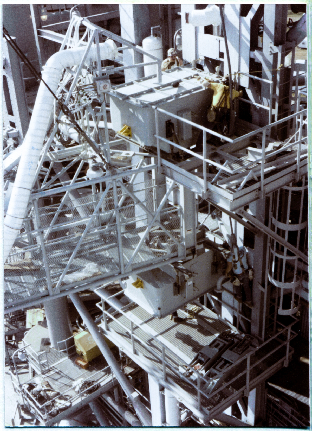 Image 136. OAA Lift 8 of 9. The laborious and time-consuming process of finish-bolting the Orbiter Access Arm to Side 1 of the FSS at Space Shuttle Launch Complex 39-B continues. I have departed the Cable Tray Access Catwalk, where Image 135, of Wade and Kevin Ivey, was taken, gone back across that Catwalk and down its Access Ladder to FSS Elevation 200'-0”, around to the other side of the FSS and up one level of stairs, to Elevation 220'-0”, and from there out to the northeast corner of the FSS and then on to the end of the top level of the Intertank Access Arm, not quite twenty full feet, eye-level, above the Upper Truss Element of the OAA, where I turned around, looked back across and down at the work in progress, and took this frame. Union Ironworkers working for Ivey Steel are hard at it, continuing to guide the crane operator who continues to fine-reposition the Arm with a surgeon's precise touch, still using pins, bars, and that uncanny ironworker-combination of overwhelming brute-force and delicate finesse, to get every last one of the bolt-hole sets, half on the Tower and their mating half on the Arm, into PERFECT alignment, so as that the Arm can then receive its final bolts and torque-down, get looked at good and close one last time, and then the crane can be unhooked from the Arm, which is now fully self-supporting, cantilevered over sixty feet out and away from the FSS nearly 200 feet above the bottom of the Flame Trench. There is, of course, no margin for error with any of this work. None at all. And if you ever find yourself accepted among those who can do it, consider yourself as having been admitted to some pretty goddamned rarified company. Photo by James MacLaren.